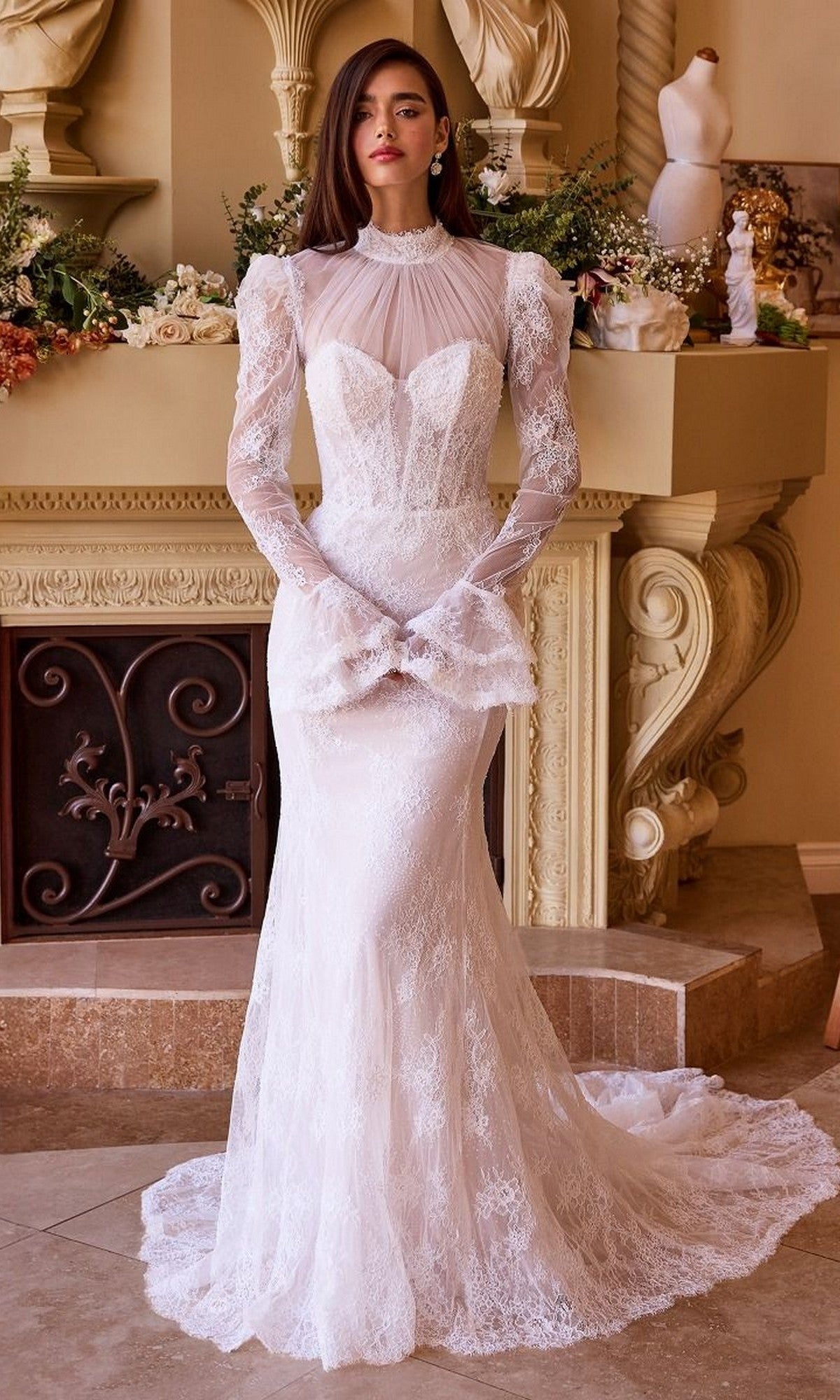 A woman stands indoors in the Andrea and Leo WL042 long wedding dress, featuring a fit-and-flare silhouette, long sleeves, white lace, high neckline, and fitted bodice, surrounded by ornate decor and floral arrangements.